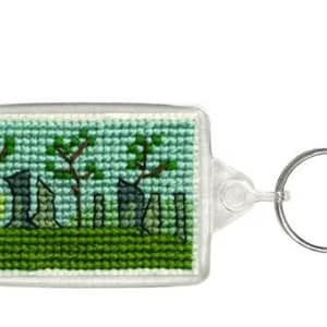 May include: A rectangular keychain with a cross-stitch design. The design features a green field, trees, and tombstones. The keychain has a clear plastic frame and a silver key ring.