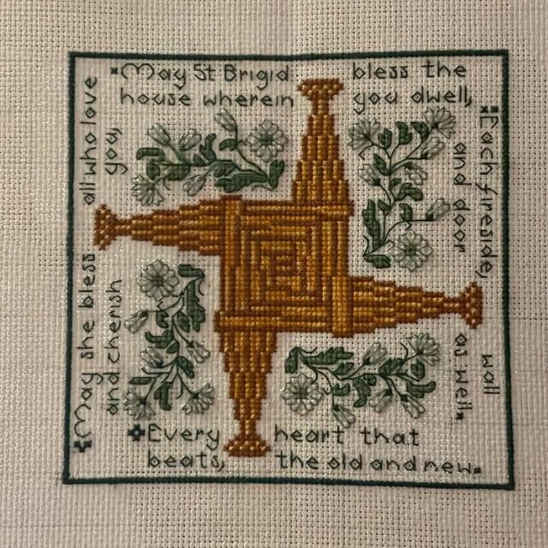 May include: A square, embroidered artwork featuring a central, golden-brown St. Brigid's cross design. Green floral accents and text surround the cross, including phrases like "May St. Brigid bless..." and "Every heart that beats..." on a cream-colored background.