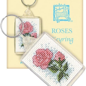 May include: Clear rectangular keychain with a cross-stitched design of pink roses and green leaves. The keychain has a silver metal ring. The background features the words "ROSES Keyring" and a blue deer logo.