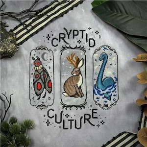 May include: A cross-stitch embroidery on light gray fabric featuring the text "CRYPTID CULTURE" and three vertical panels. The panels depict a moth with red eyes, a jackalope, and a blue sea creature in water, all surrounded by stars.