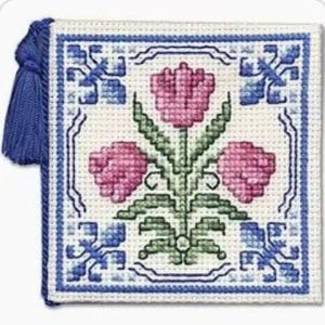 May include: Square, cross-stitched textile featuring a floral design with three pink tulips and green leaves. The border is blue and white, with blue corner accents. A blue tassel hangs from the side. The textile is likely a decorative item.