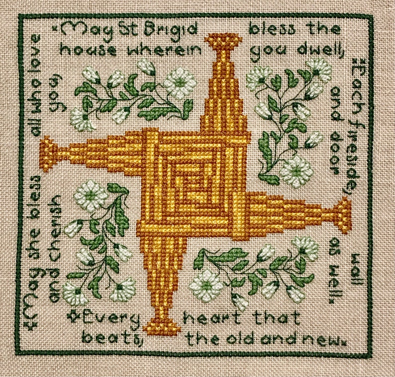 May include: A cross-stitch design featuring a central woven structure in shades of gold and brown, surrounded by floral accents and text. The text reads "May St Brigid bless the house wherein you dwell, each fireside and door as well, and cherish all who love you, every heart that beats the old and new."