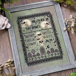 May include: Cross-stitch artwork in a grey wooden frame. The design features skulls, tombstones, and the text "Hallowed Be Thy Bones." The colour palette includes black, white, and shades of green. The artwork has a spooky, gothic theme.