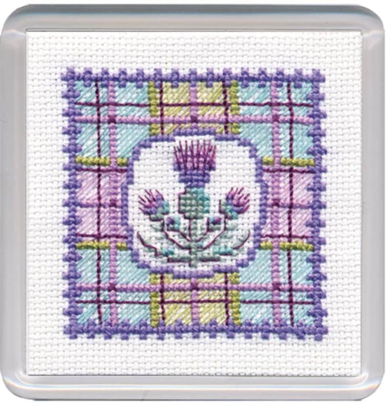 May include: A square, cross-stitched design featuring a thistle in a white oval, set against a plaid pattern in shades of blue, purple, and yellow. The design is encased in a clear, square frame.
