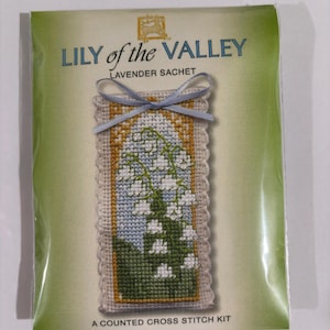 May include: A counted cross stitch kit featuring a lavender sachet with a Lily of the Valley design. The sachet is rectangular with a scalloped edge and a blue ribbon. The packaging is green and cream-colored, with text that reads "LILY of the VALLEY".
