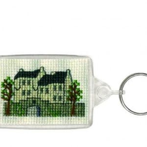May include: A rectangular keyring featuring a cross-stitch design of a white building with a black roof, green trees, and a small figure. The keyring has a clear plastic casing and a silver metal ring.