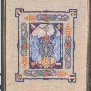 May include: Framed cross-stitch artwork featuring a mythical dragon with blue wings and a fiery breath. The design includes Celtic knot borders and corner emblems, all in a variety of colors, framed in a black wooden frame.