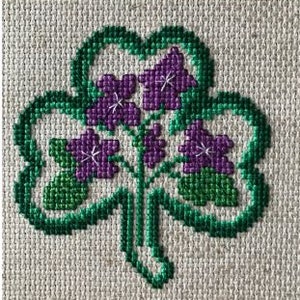 May include: A green shamrock with purple flowers embroidered on a white fabric background.