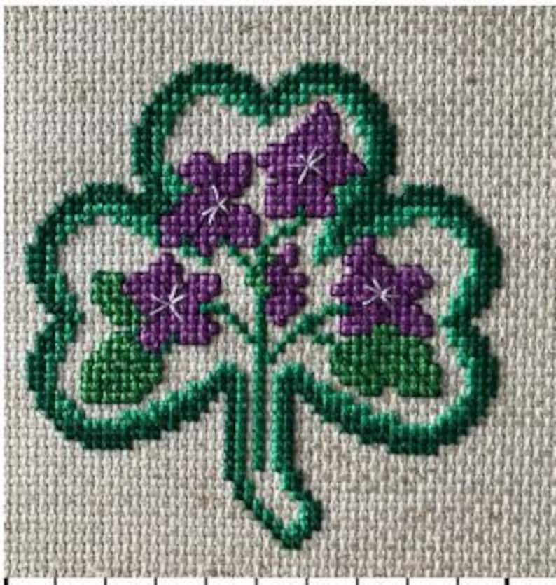 May include: A green shamrock with purple flowers embroidered on a white fabric background.