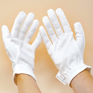 Pode incluir: Um par de luvas brancas de algod&atilde;o com uma etiqueta que diz "THESE HANDS LOTION". As luvas s&atilde;o feitas de um tecido macio e texturizado e t&ecirc;m um pulso ajustado. O fundo &eacute; de cor bege claro.