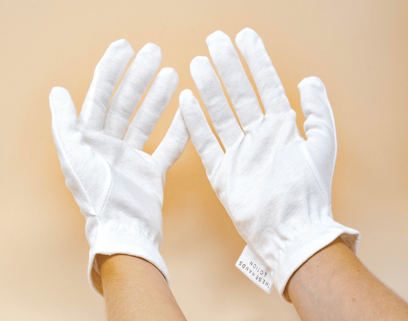 Pode incluir: Um par de luvas brancas de algod&atilde;o com uma etiqueta que diz "THESE HANDS LOTION". As luvas s&atilde;o feitas de um tecido macio e texturizado e t&ecirc;m um pulso ajustado. O fundo &eacute; de cor bege claro.
