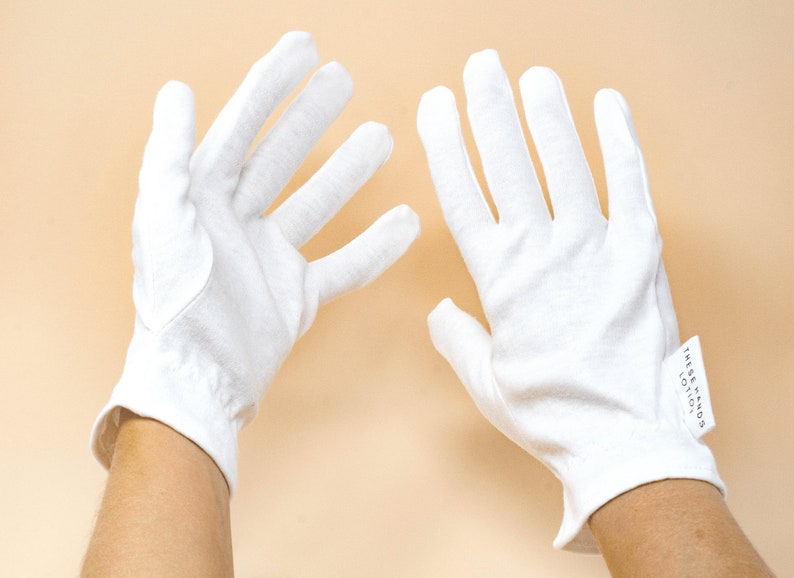 Pode incluir: Duas luvas brancas de algod&atilde;o, cada uma com punho e uma etiqueta que diz "These Hands Lotion". As luvas s&atilde;o exibidas sobre um fundo p&ecirc;ssego claro.