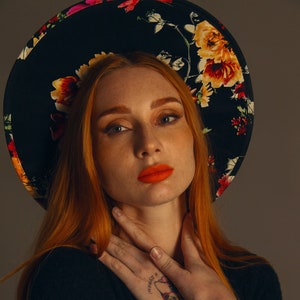 May include: A woman with long, red hair wearing a black wide-brimmed hat with floral print. The hat has a large brim and features colourful flowers. She is wearing a black sweater and has a tattoo on her hand.
