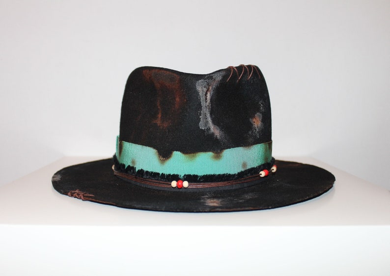 Op de afbeelding: Een zwarte wollen fedora hoed met een turquoise lint en bruine leren afwerking. De hoed heeft een verweerde look met witte en bruine markeringen. De rand is breed en de kroon is hoog.
