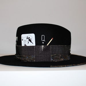 May include: Black felt fedora hat with a black and white playing card design and a matchstick on the side. The card is the Queen of Hearts.