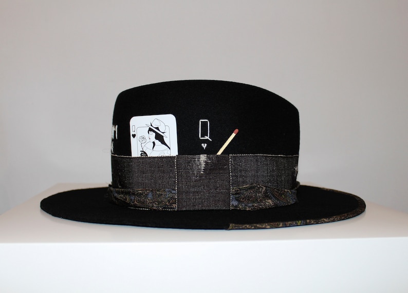 May include: Black felt fedora hat with a black and white playing card design and a matchstick on the side. The card is the Queen of Hearts.
