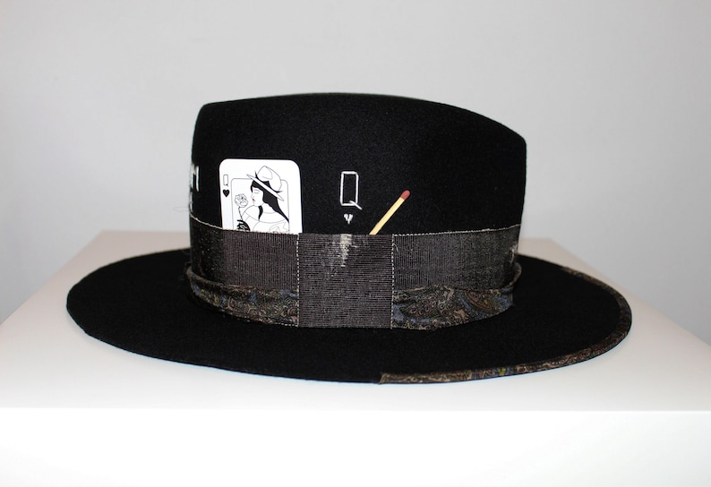 May include: A black felt fedora hat with a black and white playing card of the Queen of Hearts and a matchstick attached to the band.