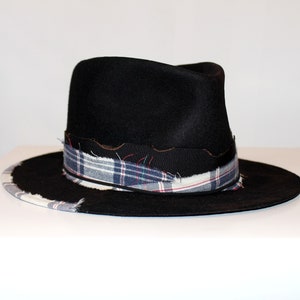 Puede incluir: Un sombrero fedora de fieltro negro con ala ancha. El sombrero tiene una banda de tela a cuadros con bordes deshilachados.