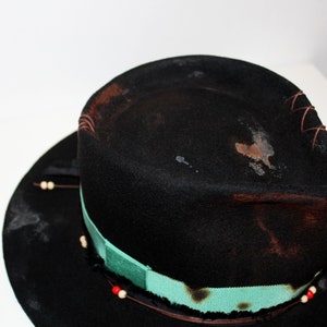Op de afbeelding: Een zwarte wollen fedora hoed met een turquoise lint en witte kralen rond de rand. De hoed heeft een verweerde look met bruine vlekken en stiksels.