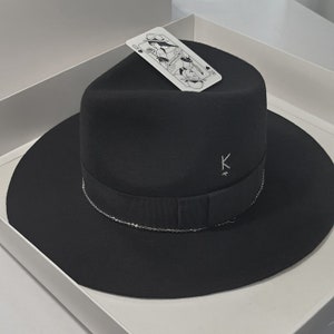 Puede incluir: Un sombrero fedora negro con una cinta negra y un detalle de cadena plateada. El sombrero tiene un logotipo 'K' en la parte delantera.