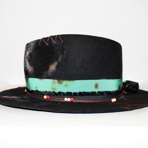 Op de afbeelding: Een zwarte wollen fedora hoed met een turquoise lint, een bruin leren koord en rode en witte kralen. De hoed heeft een verweerde look met bruine vlekken en stiksels.