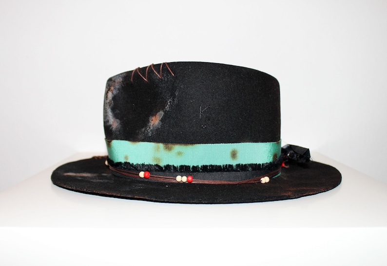 Op de afbeelding: Een zwarte wollen fedora hoed met een turquoise lint, een bruin leren koord en rode en witte kralen. De hoed heeft een verweerde look met bruine vlekken en stiksels.