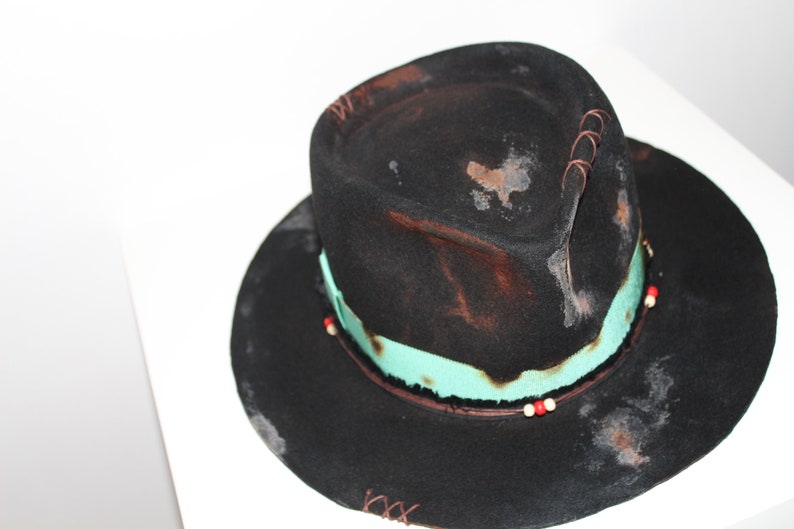 Op de afbeelding: Een zwarte wollen fedora hoed met een turquoise band en rode en witte kralen. De hoed heeft een verweerde look met bruine en witte vlekken.