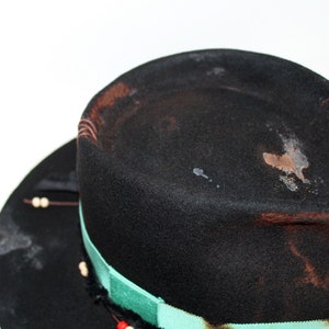 Op de afbeelding: Een zwarte wollen fedora hoed met een groen-wit lint. De hoed heeft een verweerde look met bruine vlekken en stiksels.