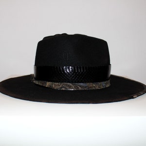 Puede incluir: Un sombrero fedora negro de ala ancha y una banda de cuero negro con estampado de piel de serpiente. El sombrero tiene una banda estampada en negro y dorado debajo de la banda de cuero.