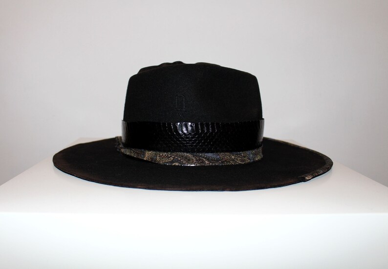 Puede incluir: Un sombrero fedora negro de ala ancha y una banda de cuero negro con estampado de piel de serpiente. El sombrero tiene una banda estampada en negro y dorado debajo de la banda de cuero.
