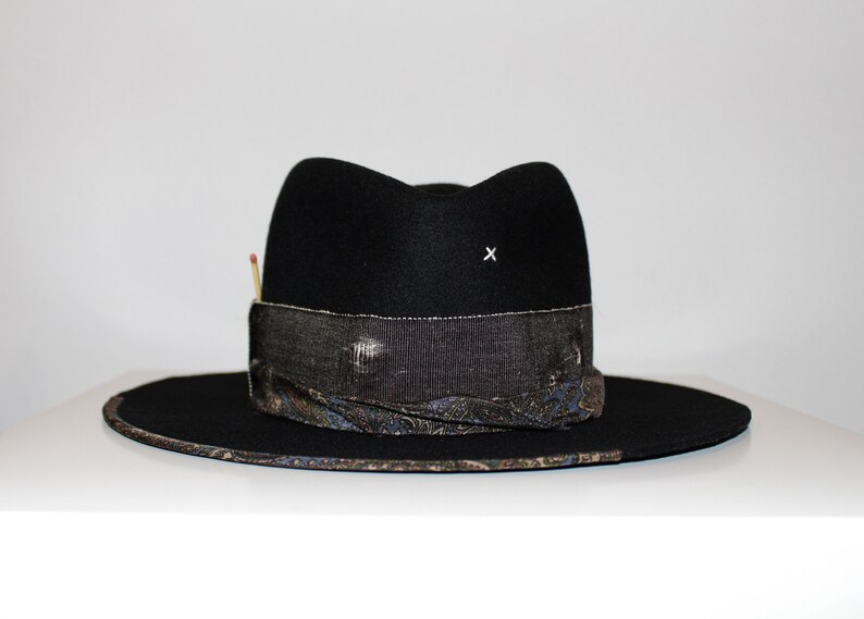 May include: A black felt fedora hat with a wide brim and a patterned band. The hat has a small white 'x' on the crown.