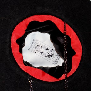 Puede incluir: Sombrero de fieltro negro con una cinta roja alrededor del borde. El interior del sombrero est&aacute; forrado con tela blanca y presenta una ilustraci&oacute;n en blanco y negro de una carta de juego con el texto "Little Red" y "I Rule The World". Una cadena negra con un broche cuelga del interior del sombrero.