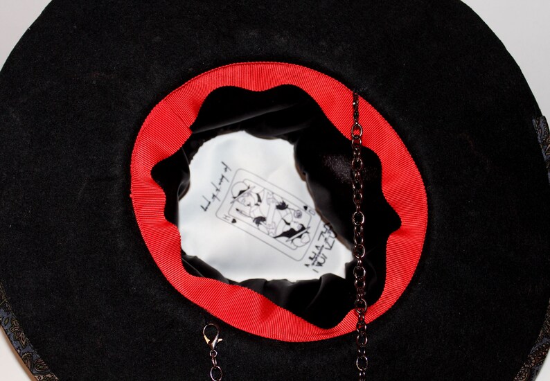 Puede incluir: Sombrero de fieltro negro con una cinta roja alrededor del borde. El interior del sombrero est&aacute; forrado con tela blanca y presenta una ilustraci&oacute;n en blanco y negro de una carta de juego con el texto "Little Red" y "I Rule The World". Una cadena negra con un broche cuelga del interior del sombrero.