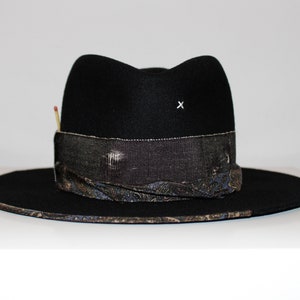 May include: A black felt fedora hat with a patterned band and a small white 'X' on the crown. The hat has a wide brim and a slightly indented crown.