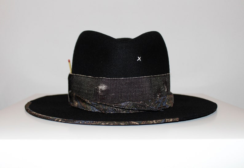 May include: A black felt fedora hat with a patterned band and a small white 'X' on the crown. The hat has a wide brim and a slightly indented crown.