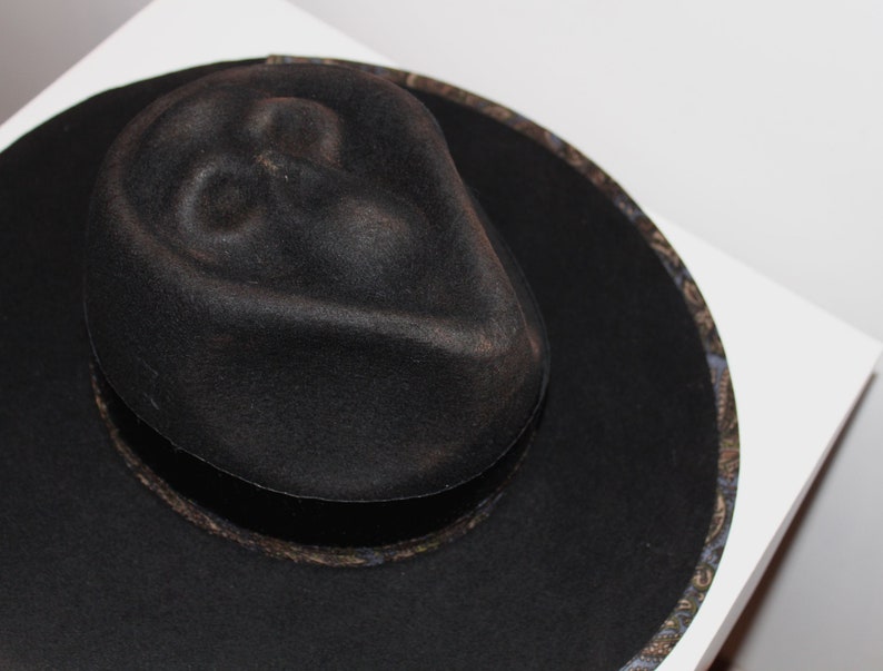 Puede incluir: Un sombrero fedora de fieltro negro con una banda estampada. El sombrero tiene una ala ancha y una copa ligeramente hundida.