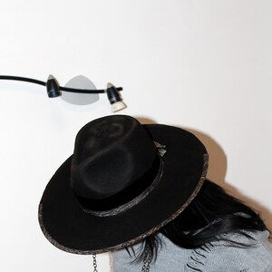 Puede incluir: Un sombrero fedora negro con una banda estampada y una cadena adjunta. El sombrero lo lleva una persona con el pelo largo y negro y un su&eacute;ter gris.