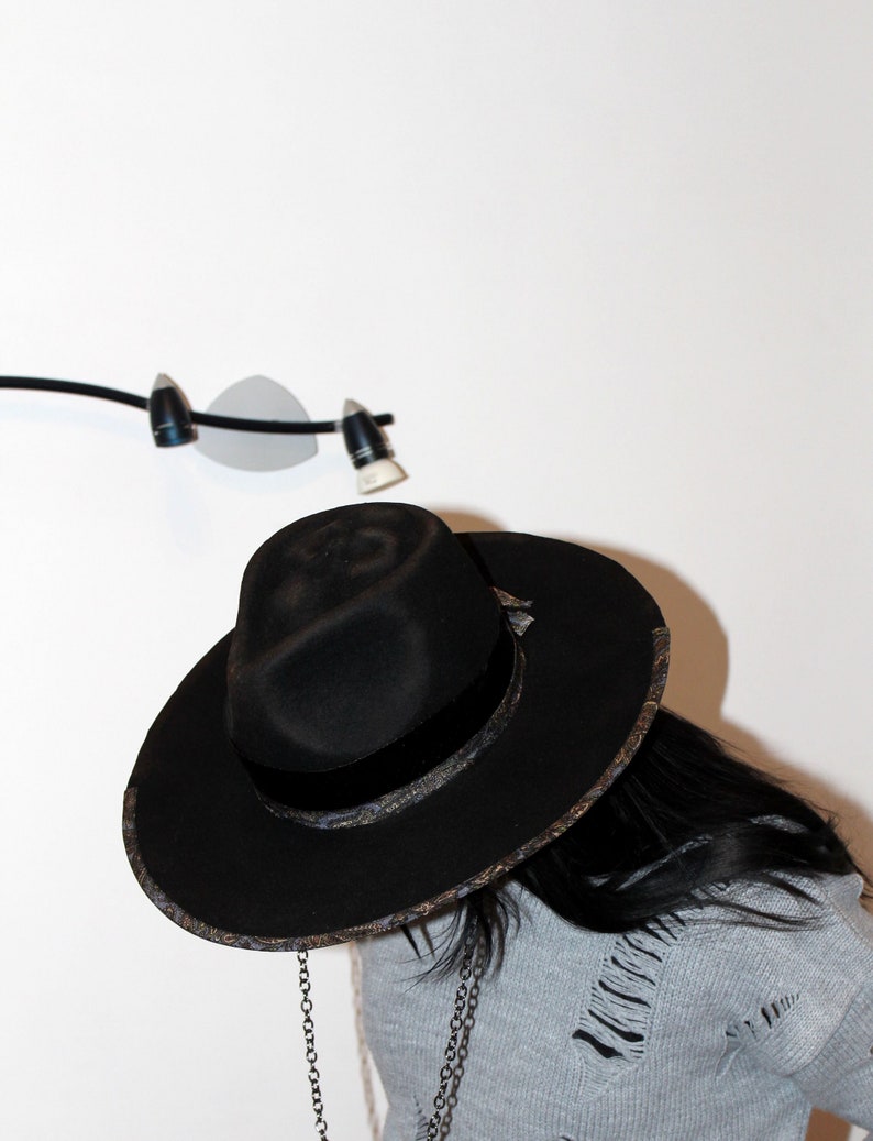 Puede incluir: Un sombrero fedora negro con una banda estampada y una cadena adjunta. El sombrero lo lleva una persona con el pelo largo y negro y un su&eacute;ter gris.
