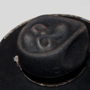 Puede incluir: Un sombrero fedora negro con una banda estampada. El sombrero tiene una corona ligeramente hundida.