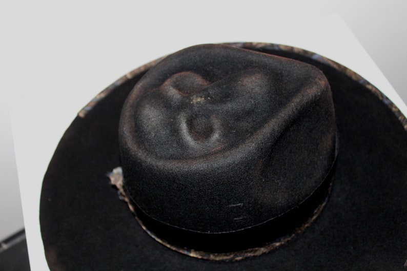 Puede incluir: Un sombrero fedora negro con una banda estampada. El sombrero tiene una corona ligeramente hundida.