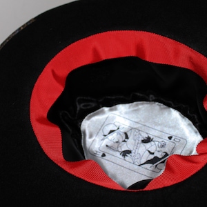 May include: Black felt fedora hat with a red ribbon around the brim. The inside of the hat has a white satin lining with a playing card design.