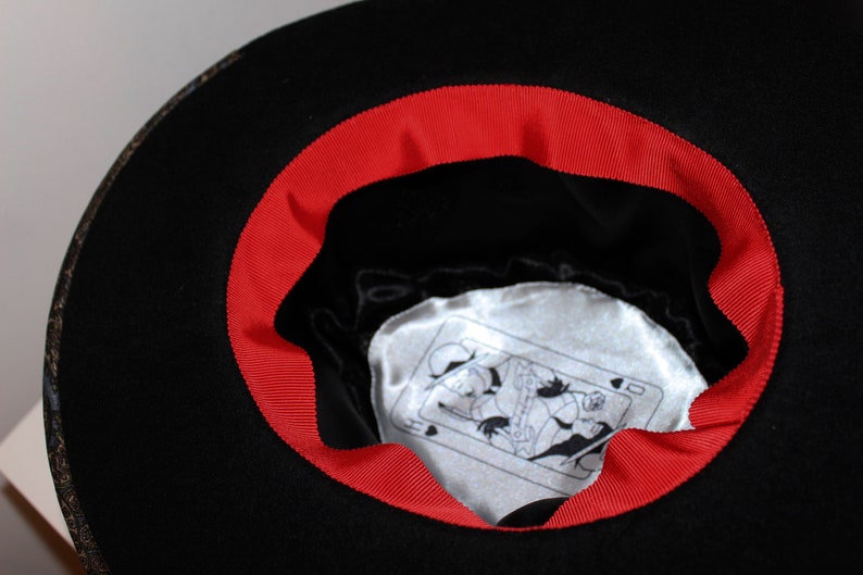 May include: Black felt fedora hat with a red ribbon around the brim. The inside of the hat has a white satin lining with a playing card design.