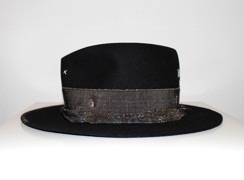 May include: A black felt fedora hat with a patterned band. The band is made of a dark blue fabric with a paisley design. The hat has a small white X stitched on the crown.