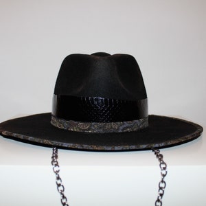 Puede incluir: Un sombrero fedora negro con una banda de cuero negro y una banda con estampado paisley debajo. El sombrero tiene una cadena.