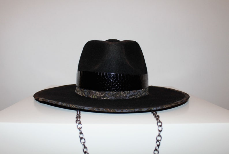 Puede incluir: Un sombrero fedora negro con una banda de cuero negro y una banda con estampado paisley debajo. El sombrero tiene una cadena.