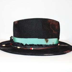 Op de afbeelding: Een zwarte wollen fedora hoed met een turquoise lint, bruine leren versiering en witte en rode kralen.