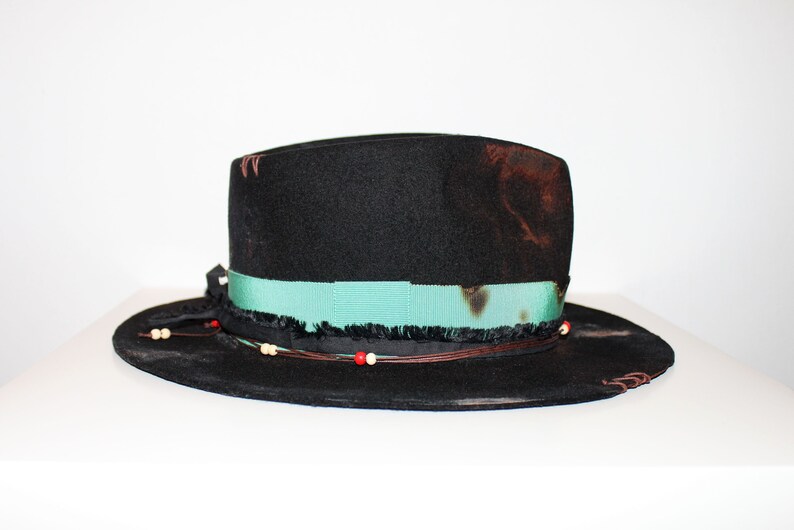 Op de afbeelding: Een zwarte wollen fedora hoed met een turquoise lint, bruine leren versiering en witte en rode kralen.