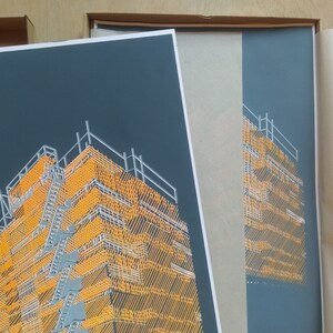May include: A close-up of a framed print of a building with a yellow and grey geometric pattern. The print is in a black frame and is being packaged for shipping.