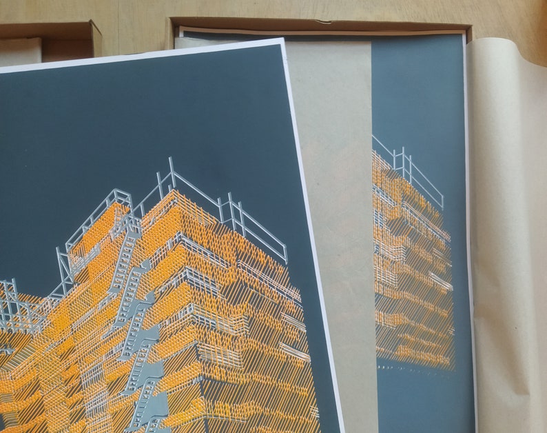 May include: A close-up of a framed print of a building with a yellow and grey geometric pattern. The print is in a black frame and is being packaged for shipping.