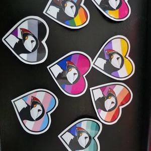 Könnte beinhalten: Neun herzförmige Aufkleber mit einem Cartoon-Papageitaucher auf jedem. Die Papageitaucher tragen alle einen roten und weißen Hut. Jeder Aufkleber zeigt eine andere Pride-Flagge, darunter die Regenbogen-, Bi-, Transgender-, Non-Binary-, Pansexual-, Lesben- und Asexual-Flaggen.